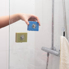 Hand holding a blue cleaning tool against a shower wall with soap bars attached.
