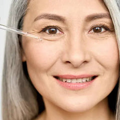A smiling woman with gray hair holding a dropper bottle of serum, with a blurred background.