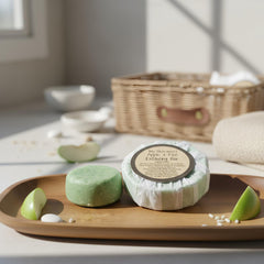 Two soap bars on a wooden tray with a basket and towel in the background.